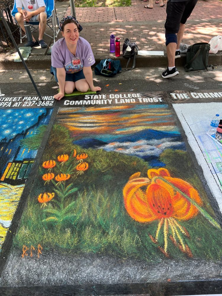 The artist poses with a large 4 foot by 6 foot chalk painting on asphalt, featuring an orange lily in the foreground with mountains, mist, and a sunset in the background.