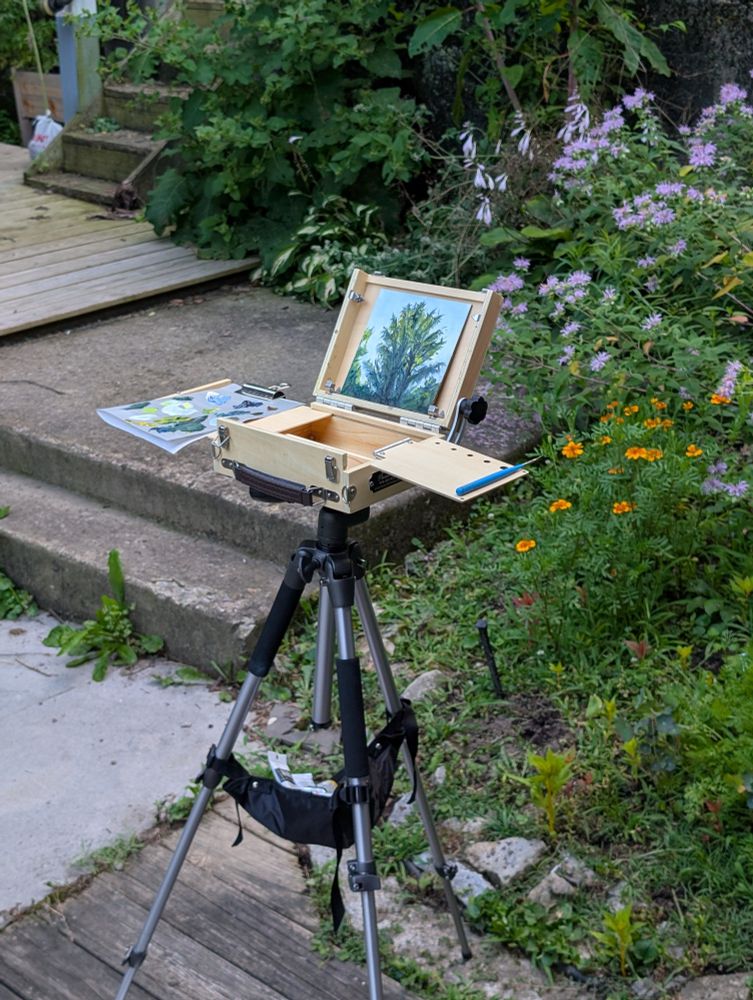 A pochade box holding a small oil painting sits on a tripod in front of a garden.