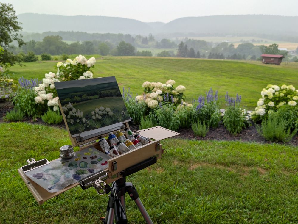A plein air oil painting set up on a tripod in front of some hydrangeas, a field, and some ridges in the background.