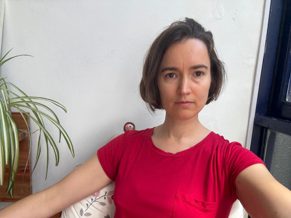 me, sitting in a chair with an embroidered pillow behind me; spider plant leaves fall over a shelf to my right and a blue window frame can be seen to my left. Also, a small puppet head with magenta hair, a green cap and a goofy expression is peeking over my right shoulder.