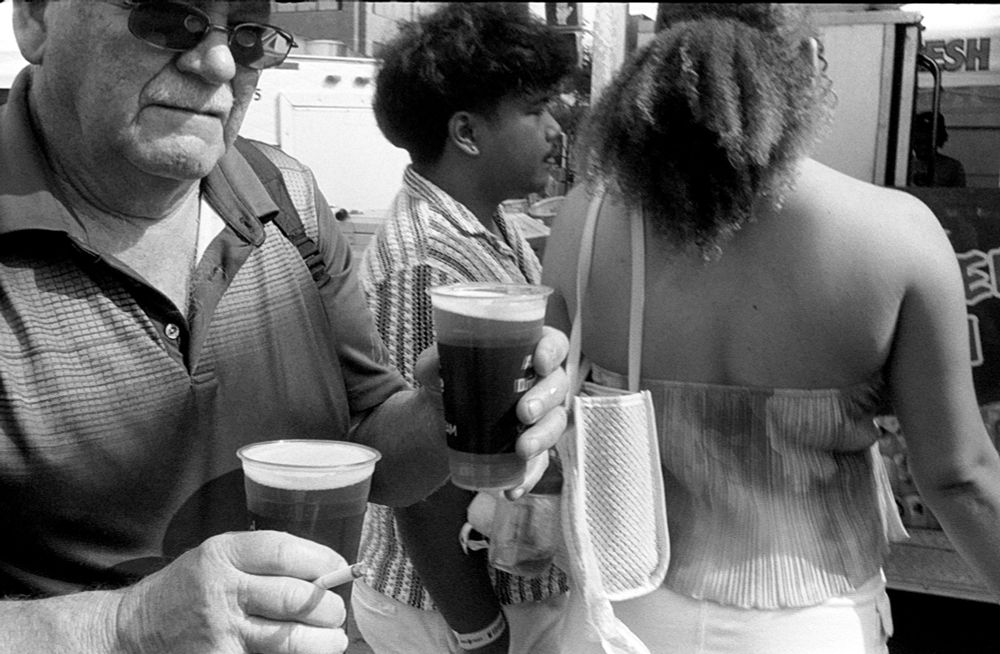 Black and white photo of a man at a street fair, holding a beer in each hand with a cigarette in between the first two fingers of his right hand Behind him a young couple orders a drink