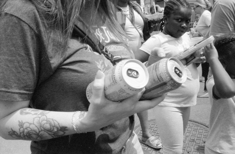 Black and white photo of a crowd. In the close foreground a woman who’s head is cut off by the top of the frame is holding two large cans in one hand. Behind her a young girl is looking at the camera 