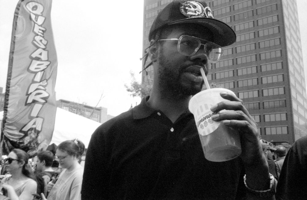 Black and White photo of a black man in a black polo, hat, and cool looking clear framed glasses. He is drinking a lemonade and on the left side of the frame is a large banner reading “quesabiria”