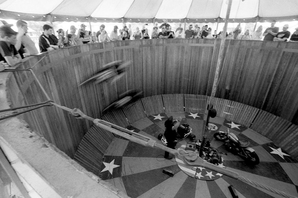 Black and white photograph of the “Wall of Death”, a large circular wooden structure with a crowd of people standing at the top. Two stunt performers are riding motorcycles along the wall of the structure and on the bottom a man is standing near another motorcycle and a go kart 