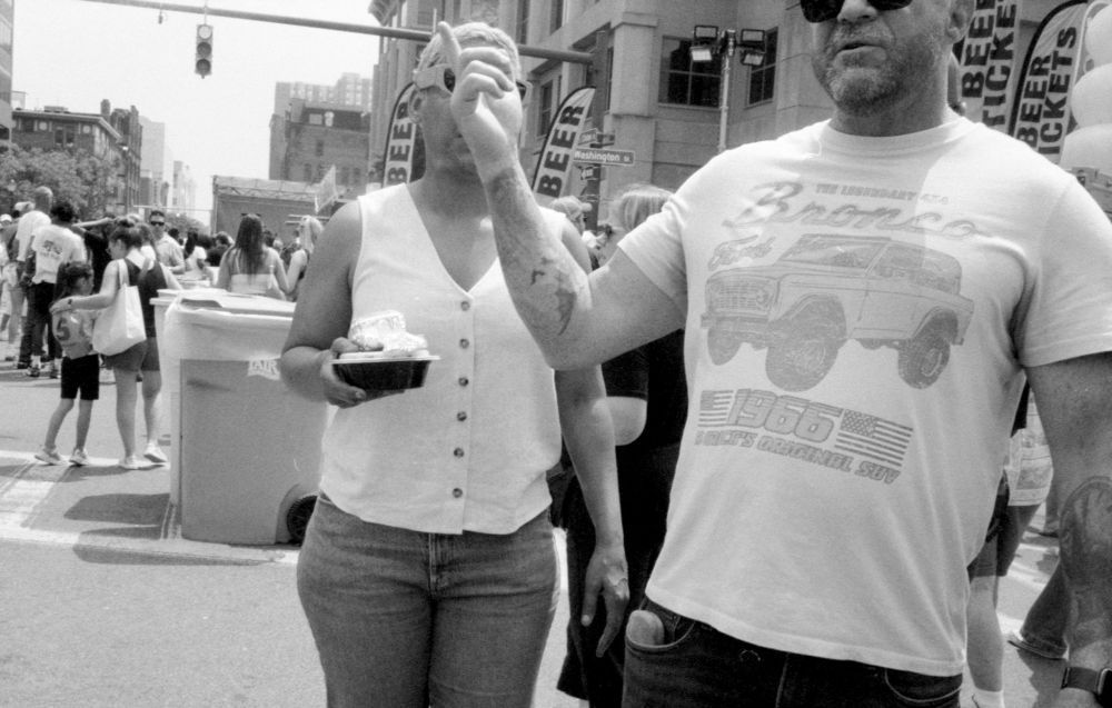 Black and white photo of a man and a woman at a street fair. The man is wearing a white Ford Bronco t shirt and is holding is right hand up pointing towards the sky. His hand is directly in front of the woman’s face, obscuring it