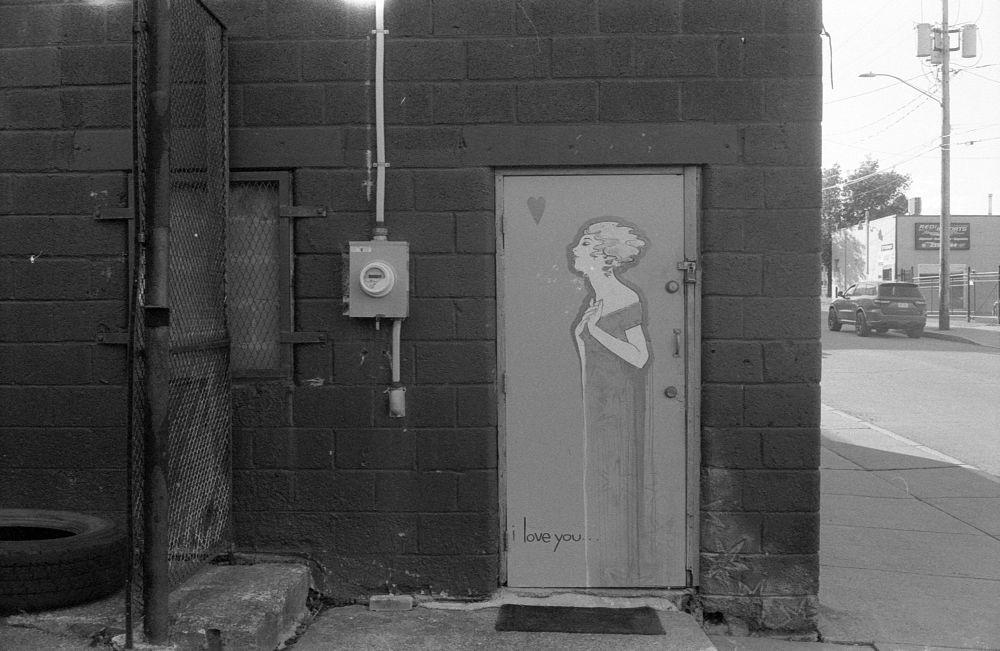 Black and white photo of a doorway in a concrete wall. The street beyond is visible on the right hand side of the frame. On the door is an art nouveau style painting of a woman with a small heart above her head. At the bottom of the door “I Love You” is written in small letters 