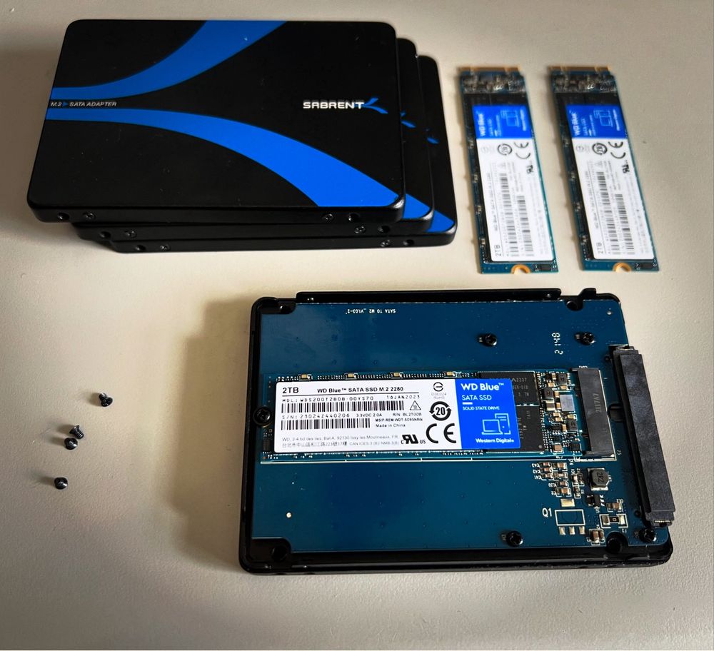 A collection of computer storage devices, including stacked Sabrent M.2 SATA adapters, with WD Blue SATA SSD M.2 drives, an opened SSD enclosure, and a few small screws on a light-colored surface.
