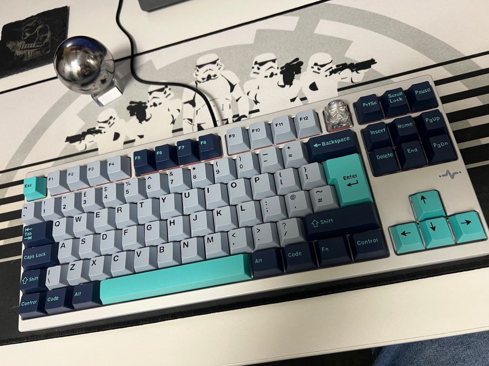 An overhead view of a mechanical keyboard with a turquoise and gray color scheme. The keyboard features a custom stormtrooper keycap and is placed on a surface with a design of stormtroopers in the background.