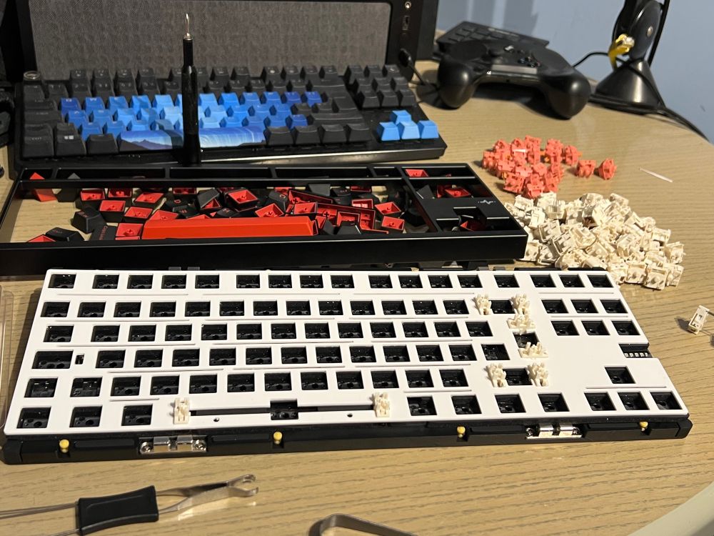 An assortment of keyboard components is displayed on a desk. A disassembled keyboard base with some key switches installed sits in the center, while various colored keycaps and switches are arranged nearby. Additional tools are present for assembling the keyboard.