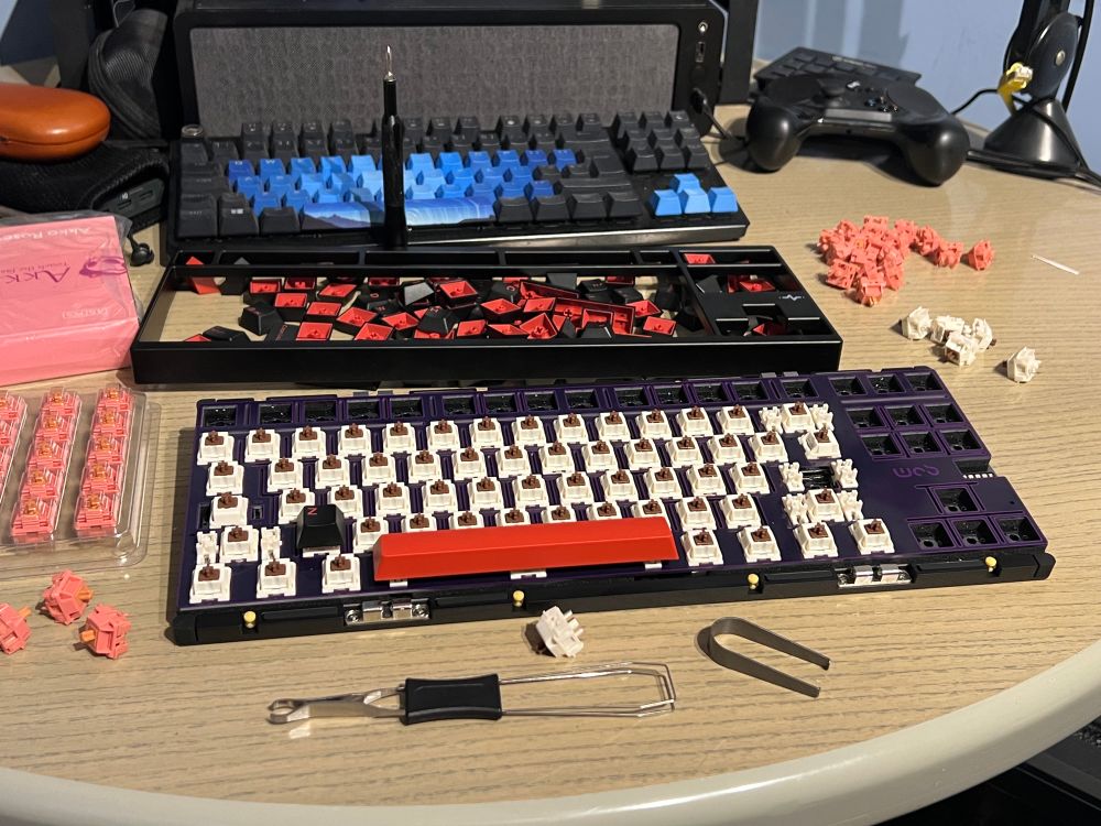 A dismantled mechanical keyboard is displayed on a table, surrounded by various key switches, keycaps, and tools for assembly. There are two keyboards in the background, one with blue keycaps and one that is partially disassembled.