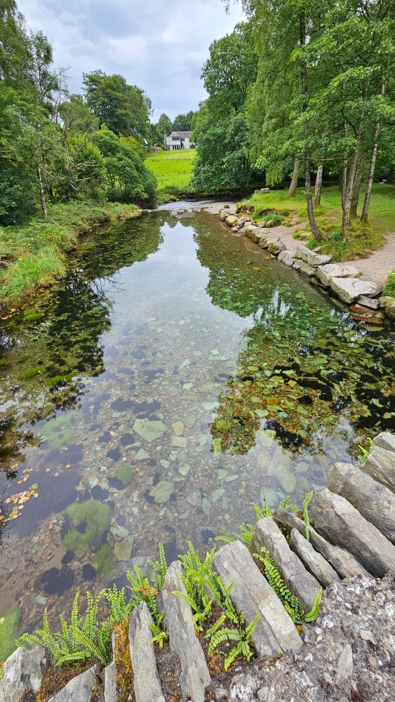 A crystal clear river with stones in the bottom. Green banks, a bridge and a house on a hill.