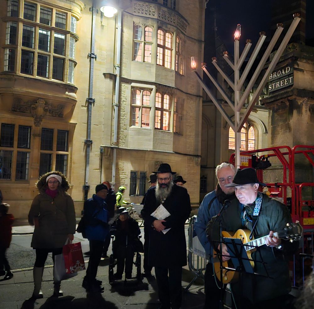 A large Chanukiah with the shammas and a single candle lit for the first night of Chanukah, in Broad St in Oxford. A man with a guitar is singing Oseh shalom.