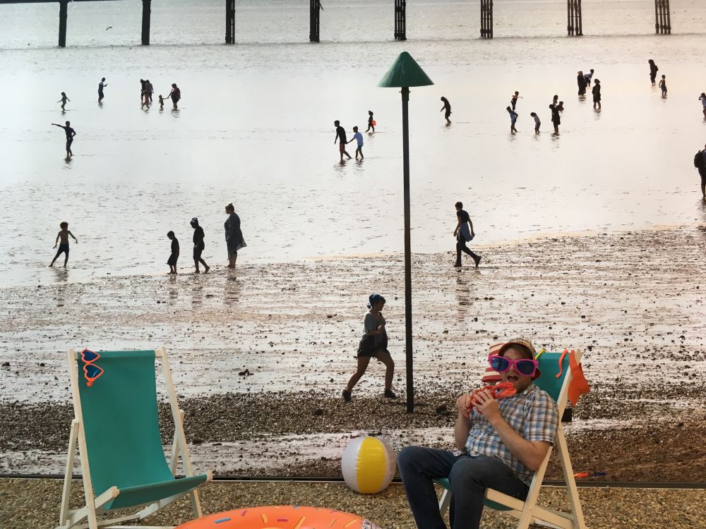Silhouetted figures on a beach. It's a huge print. In front I am sitting on a deck chair, wearing outsize sunglasses and pretending to eat a whole lobster. Photo by my daughter. National Maritime Museum, Greenwich.