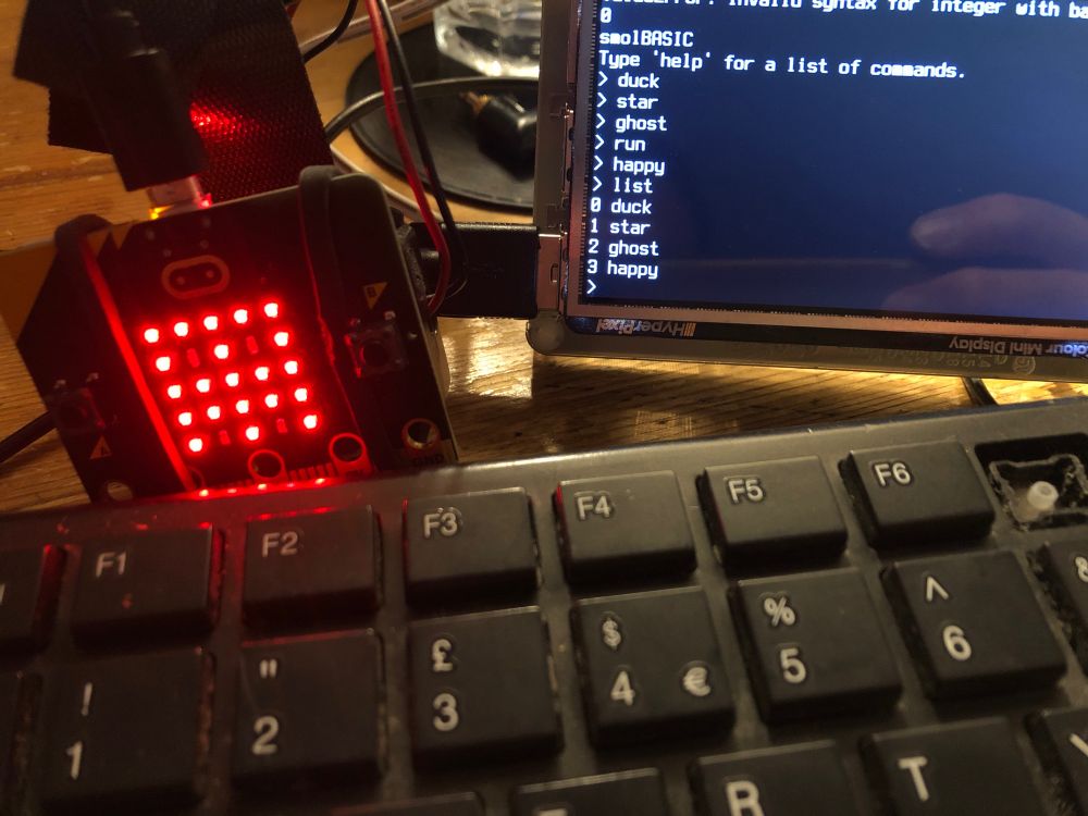 A BBC micro:bit showing a ghost icon. A USB keyboard. A small LCD display showing a program being entered, run on the micro:bit and listed. The program is:
duck
star
ghost 
happy