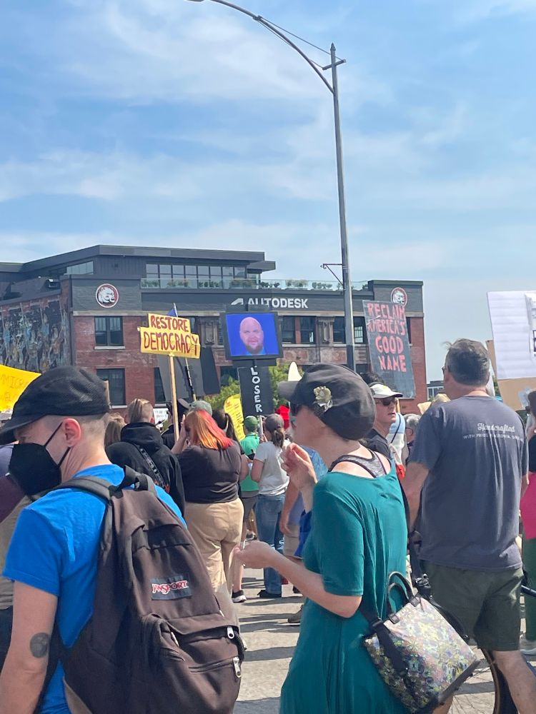Picture from the Labor Day protest in Portland, OR, many people are marching across a bridge holding signs, one of the signs is just an image of the weird bald JD Vance meme with a blue background 