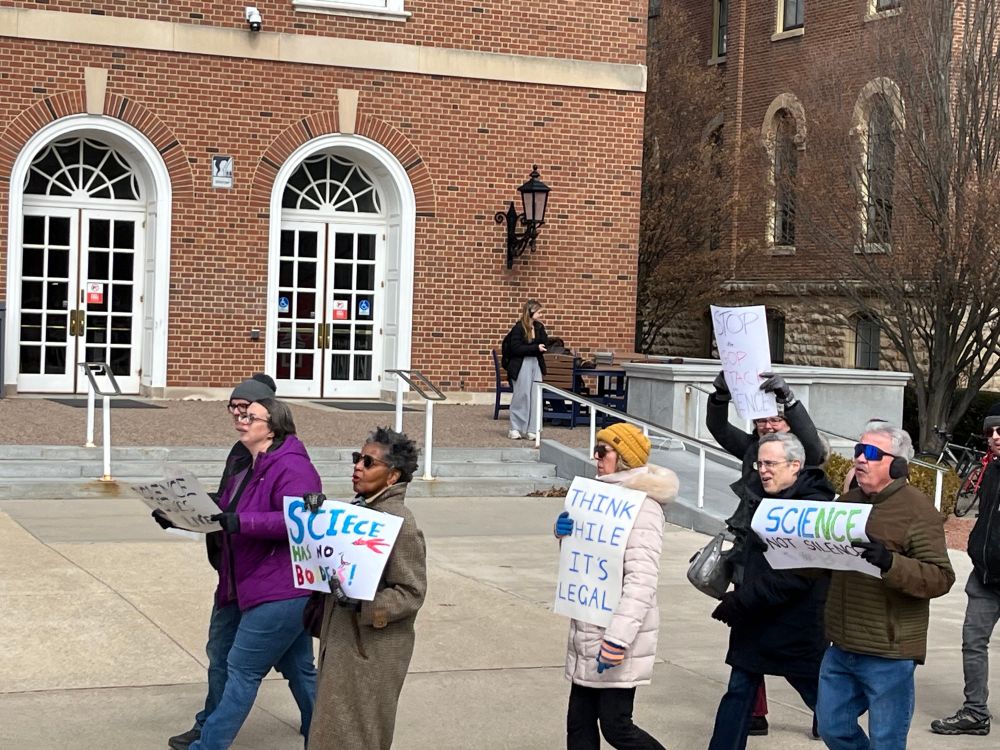 A group of people marches outside a red-brick building, holding protest signs. One sign reads, “SCIENCE has no borders,” another says, “THINK WHILE IT’S LEGAL,” and another says, “STOP ATTACKING SCIENCE.” A tall English professor holds a sign reading “Stop the GOP Attack on Science.” The protesters wear winter coats, gloves, and hats. The background shows a historic building with large white doors, arched windows, and steps leading to an entrance.