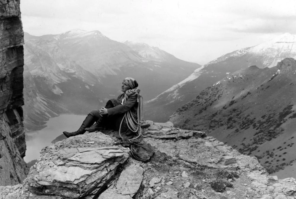 Photograph at Glacier National Park taken by Ray Bell, showing Dorothy Pilley sitting with her climbing rope on the Ptarmigan Wall, over Elizabeth Lake. Image from https://glorioussport.com/articles/dorothy-pilley-climbing-pinnacle-club-memoir/