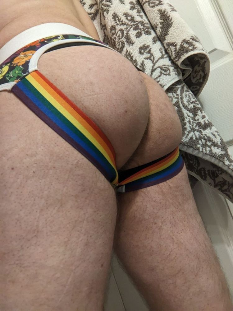 My crotch, wearing a Rudderbutts brand jockstrap with the "otterama" pattern that has rainbow colored cartoon otters printed and rainbow straps. The black straps of my chastity belt can also be seen under the jockstrap. There is a patterned towel in the background.
