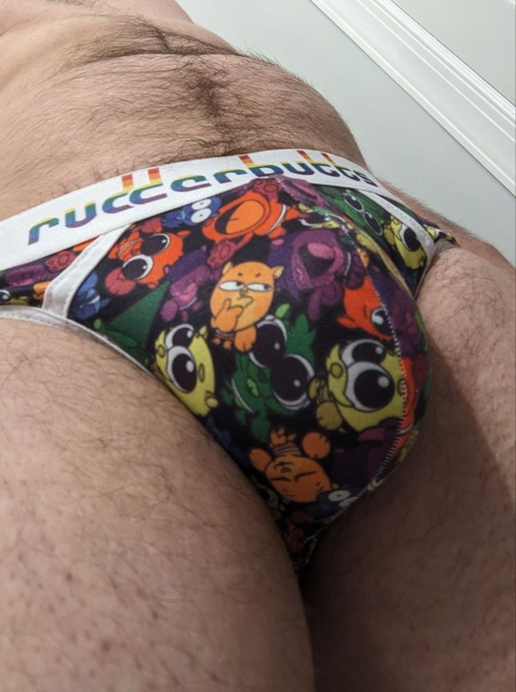My crotch, wearing a Rudderbutts brand jockstrap with the "otterama" pattern that has rainbow colored cartoon otters printed. I have hair trailing from my belly button into the front of the underwear and the imprint of my chastity cage can be seen through the jock pouch.