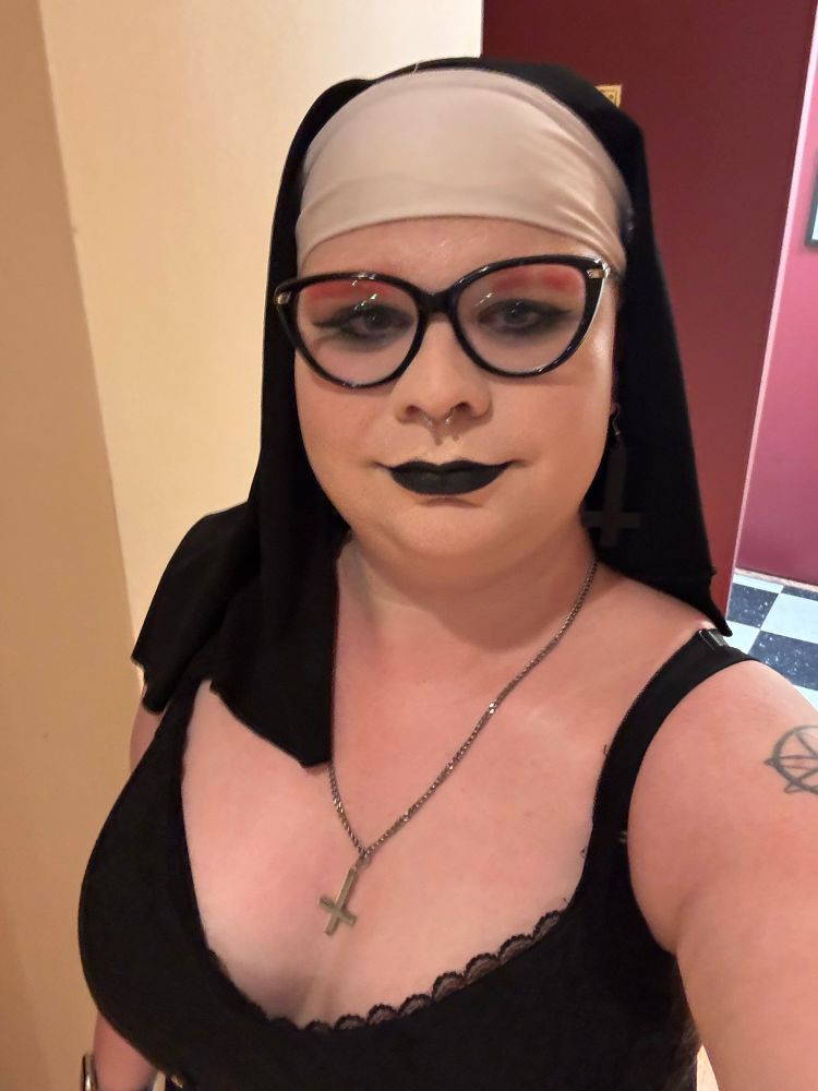 Nora is in her SistAir of Mercy stagewear, with a nun’s habit and a low cut top with an inverted cross necklace hanging between her tits.