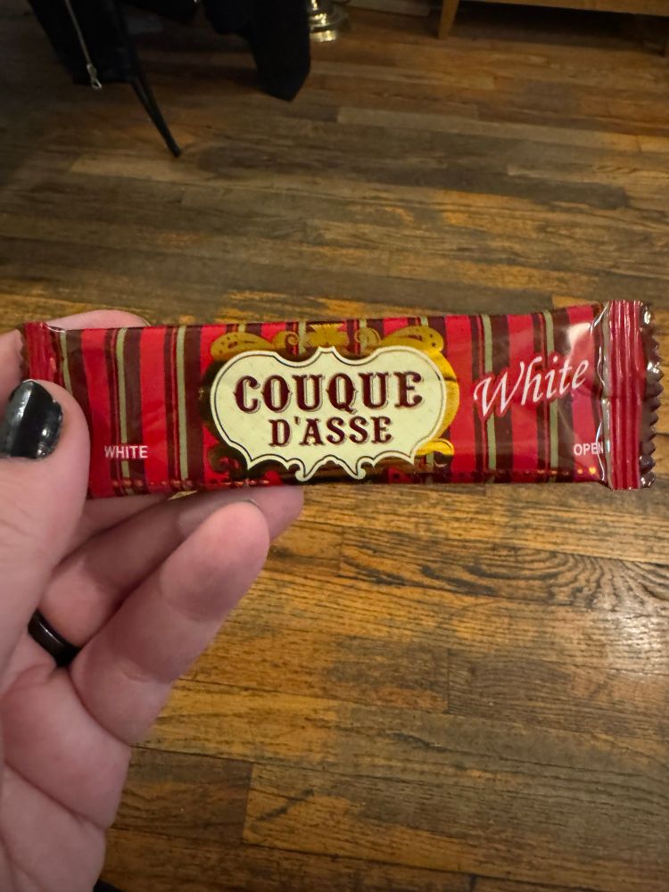 A Coque D’Asse White cookie in its wrapper. The wrapper is red with brown stripes, and the name of the cookie is in a very baroque font on an odd shaped medallion. “White” is next to it in a more contemporary looking italic serif font.