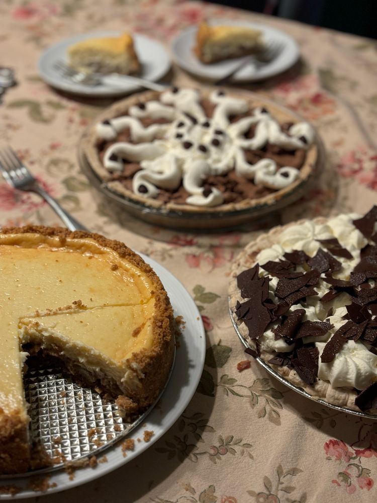 Three dessert pies on a table. They are: apple pie topped cheesecake, French silk, chocolate. 