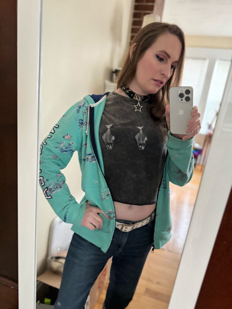 Jolene has long brown hair, pink makeup, a crop top and a blue sweatshirt over it, and jeans with a white belt