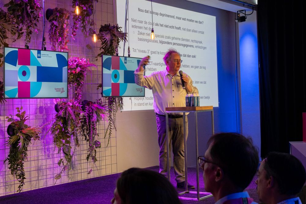 Bert Hubert op een podium met een microfoon, staand voor een scherm met daarop de tekst "Nou dat is knap deprimerend, maar wat moeten we dan?"