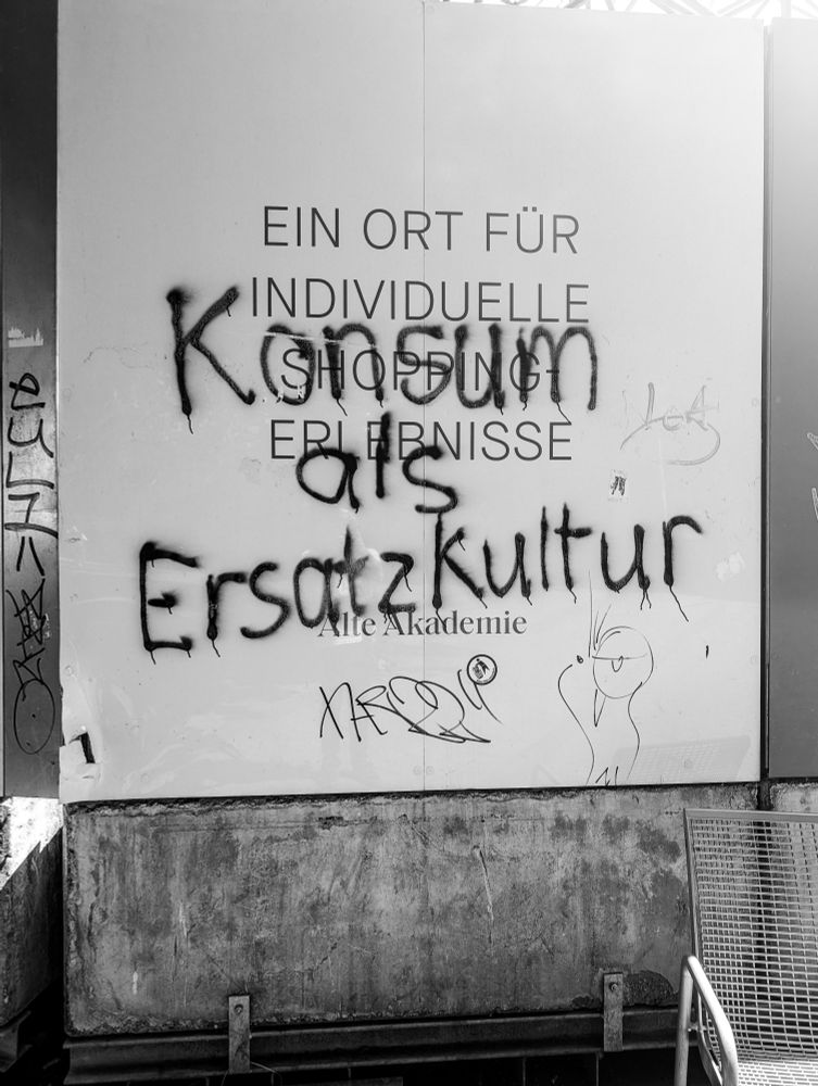 Eine große Tafel, auf welcher usprünglich stand 'Ein Ort für individuelle Shopping Erlebnisse'. Übersrprüht mit dem Spruch 'Konsum als Ersatzkultur'.