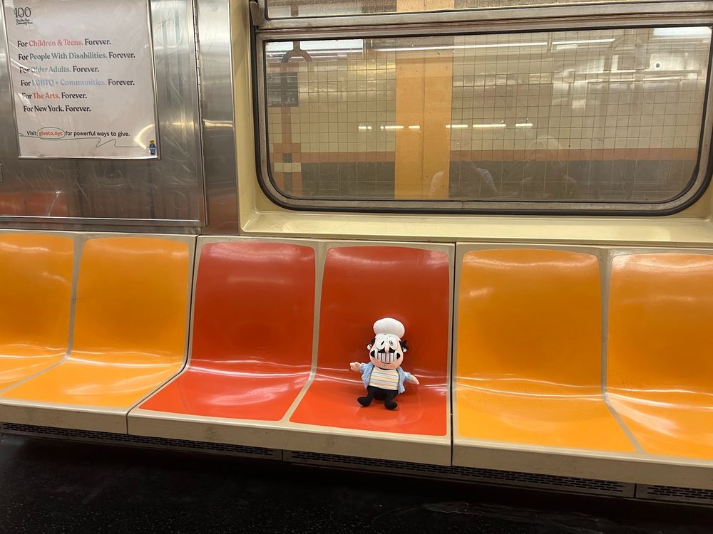 Peppino wearing a blue checkered overshirt with a multicolor striped undershirt sitting in a train on the new york subway.