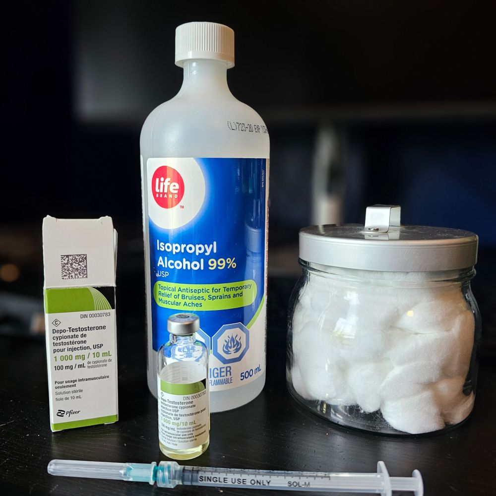 HRT still life with a jar of cotton balls, isopropyl alcohol, testosterone bottle and box, used needle and syringe. 