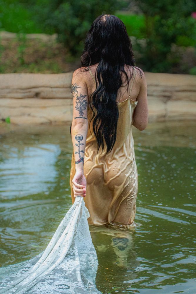 Person with long dark hair and tattoos on their arm standing waist-deep in the water at Seven Fountains Pools in Minneapolis. They are wearing a gold slip dress and holding a trailing piece of lace fabric behind them. The background features natural stone and greenery.