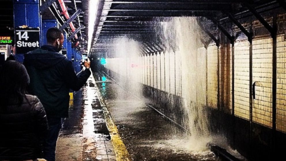 FOTO, nyc subway with water pouring down from the ceiling due to a broken water main in the street above, or similar. Someone w/ cellphone is photographing the mess. Large-text sign on a pillar says "14 Street".