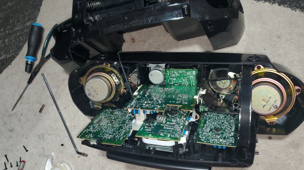 A boombox opened up to show speakers and contraptions and at least five motherboards. There are screws and tools on the floor