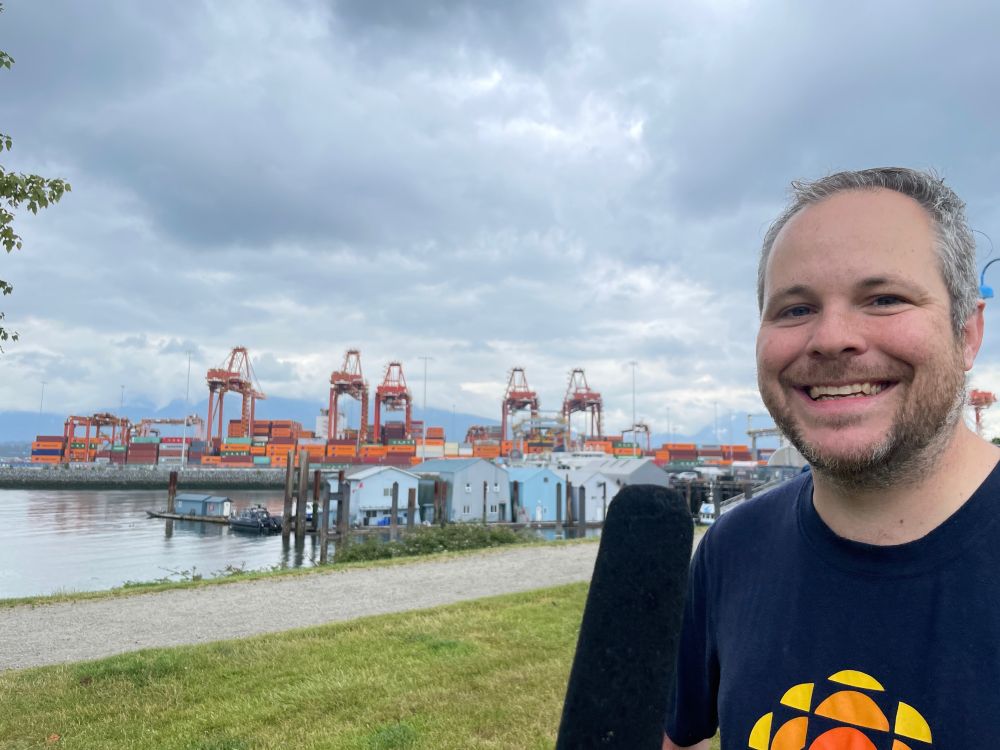 Justin McElroy has a mic stuck in his face in Vancouver's CRAB Park, on the edge of Vancouver port with stacks of shipping containers and big cranes