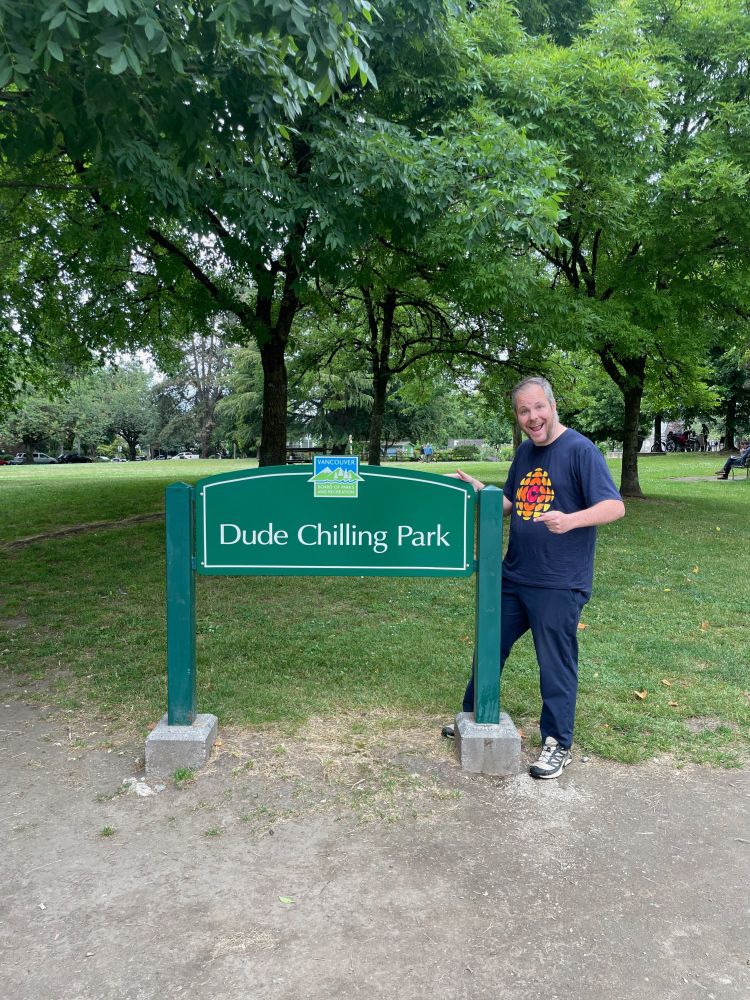 Justin McElroy poses enthusiastically with the green standing sign for Dude Chilling Park on the edge of the small park's worn grass