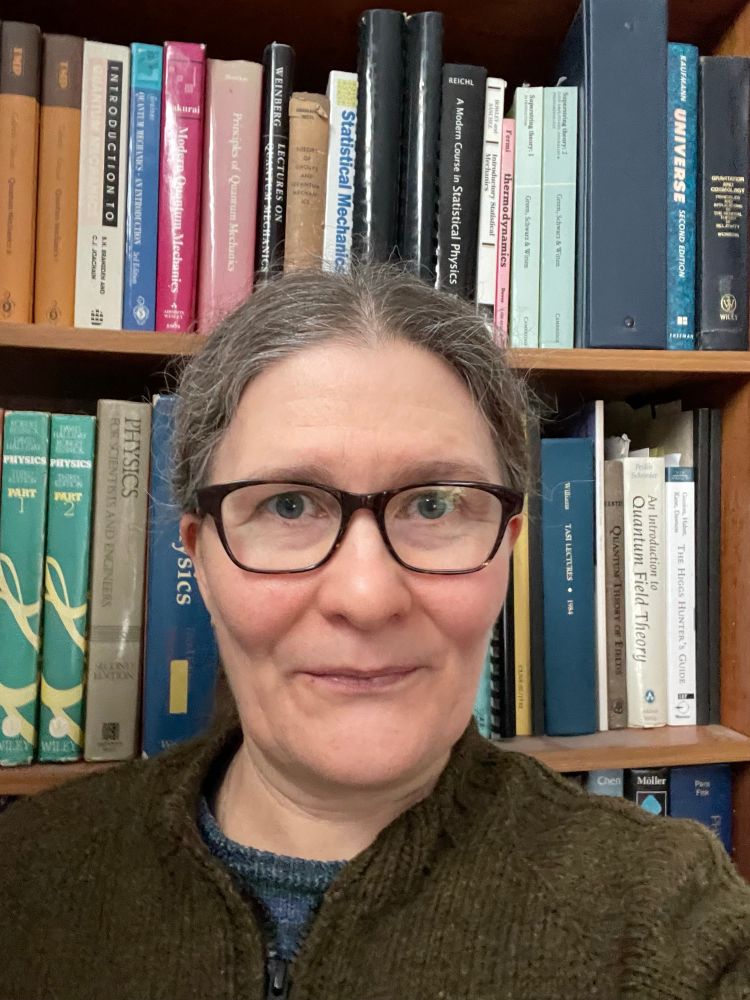 Photo of a woman with brown hair and glasses standing in front of a bookshelf, with her lips slightly pursed.