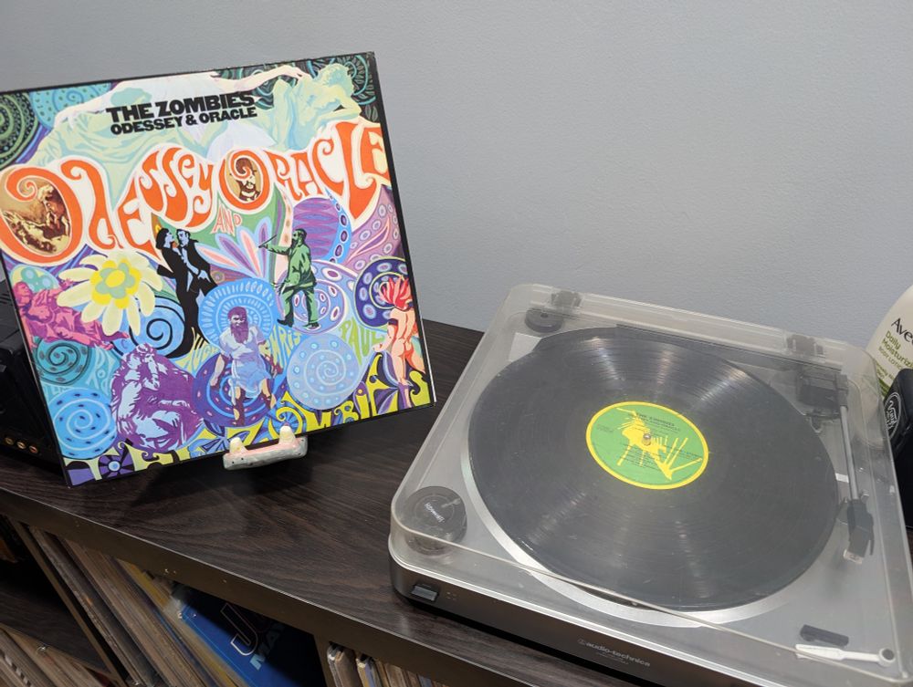 On the left is the album cover for Odessey and Oracle by the Zombies. At the top is the title and artist in black text centered on the box. Just below it is the title in psychedelic text across the cover. There are beautiful paintings of flowers and people in the psychedelic style. At the bottom is the artist name fitting into negative spaces. On the right is the album sitting on the turntable. It is waiting to be played.