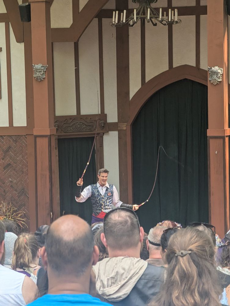 A man in a French dandy outfit and a drawn on mustache whipping in front of a crowd 