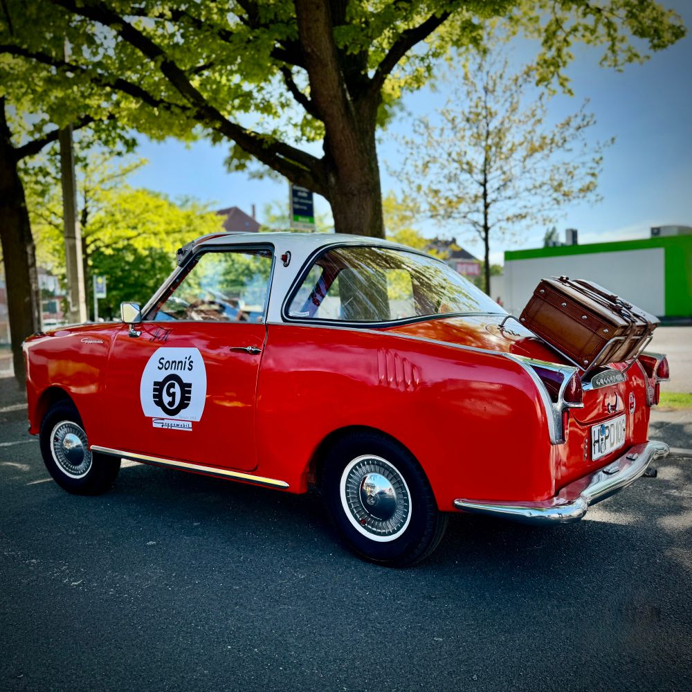 Inmitten einer ruhigen, sonnendurchfluteten Straße parkt ein kleines, leuchtend rotes Oldtimer-Auto, das sofort die Blicke auf sich zieht. Es ist ein Goggo Coupé – ein seltener und charmanter Klassiker aus den 1950er Jahren. Der Wagen glänzt frisch poliert, sein Dach ist silbern abgesetzt, und an der Rückseite ist ein brauner, nostalgischer Koffer befestigt – als wäre das Auto bereit für eine abenteuerliche Reise.

Auf der Fahrertür prangt ein rundes Emblem mit der Aufschrift „Sonni’s“ und einem stilisierten Logo. Die Szene wird von großen, üppigen Bäumen eingerahmt, deren grünes Laubspiel im Sonnenlicht tanzt. Obwohl der Wagen still auf einem unscheinbaren Parkplatz steht, strahlt er eine lebendige Energie und ein Stück vergangene Zeit aus – ein kleines, besonderes Highlight im Alltag, das Geschichten von Freiheit, Freude und Entdeckergeist erzählt.
