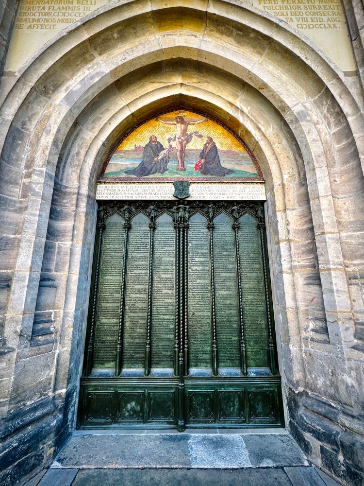 Das Foto zeigt die berühmte Tür der Schlosskirche in Wittenberg. Dort hat Martin Luther im Jahr 1517 seine 95 Thesen angeschlagen – ein wichtiger Moment der Reformation. Die Thesen sind heute in Bronze in die Tür eingraviert. Über der Tür befindet sich ein Bild von der Taufe Jesu. Die Szene spielt sich unter einem steinernen Torbogen ab.
