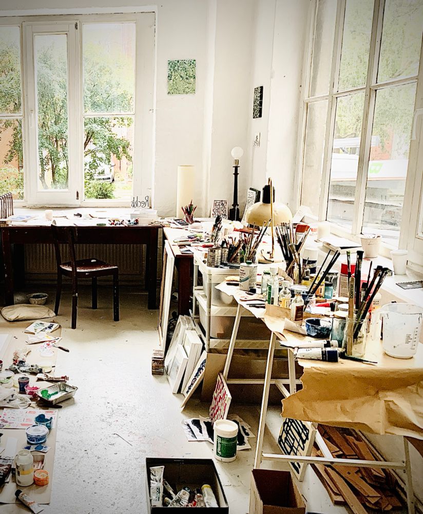 Das Foto zeigt ein helles Atelier mit großen Fenstern, durch die viel Tageslicht fällt. Der Raum ist erfüllt von „kreativem Chaos“: Überall liegen Farbtuben, Pinsel, Skizzen und Leinwände verstreut. Zwei Arbeitstische sind überfüllt mit Kunstmaterialien, und der Boden ist ebenfalls mit Malutensilien bedeckt. Trotz des Durcheinanders wirkt der Raum lebendig und inspiriert – ein Ort, an dem Kunst entsteht.