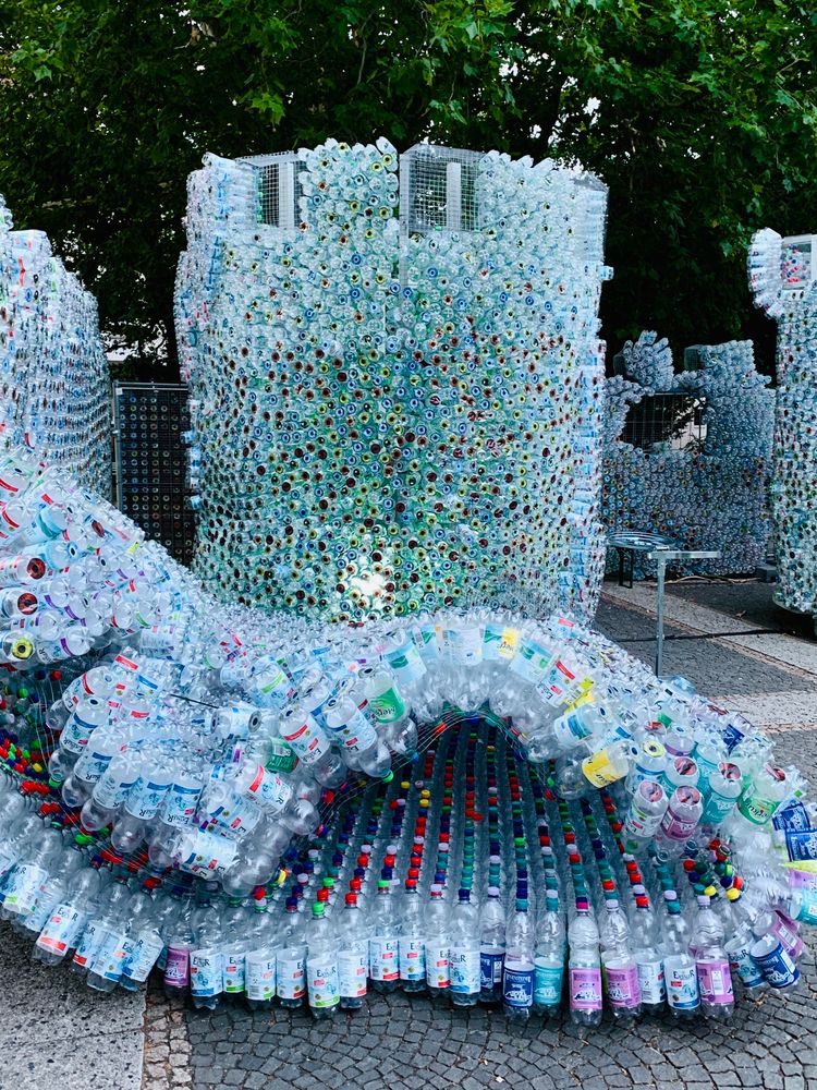 Mitten auf einem öffentlichen Platz steht eine auffällige Installation, die komplett aus leeren Plastikflaschen besteht. Mehrere hunderte, wenn nicht Tausende Flaschen sind kunstvoll aneinandergefügt – in Wellen, Türmen und Mauern. Die bunten Deckel und Etiketten leuchten in allen Farben. Die Form erinnert vage an eine Burg oder Fantasiewelt aus Müll. Es wirkt wie eine Mischung aus Mahnmal, Kunstwerk und Recyclingprojekt – laut, bunt, und irgendwie provokant.