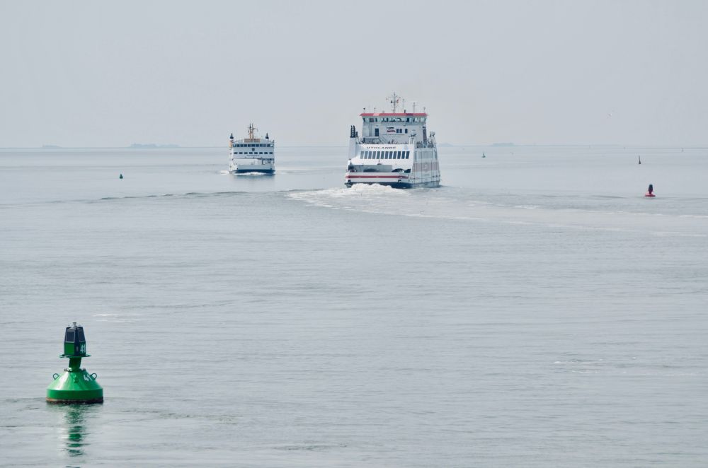 Auf ruhigem, grau-blauem Wasser fahren zwei weiße Autofähren hintereinander Richtung Horizont; ihre weißen Kielwasser zeichnen sich klar ab. Im Vordergrund schwimmt eine große grüne Fahrwassertonne als Leitmarke.