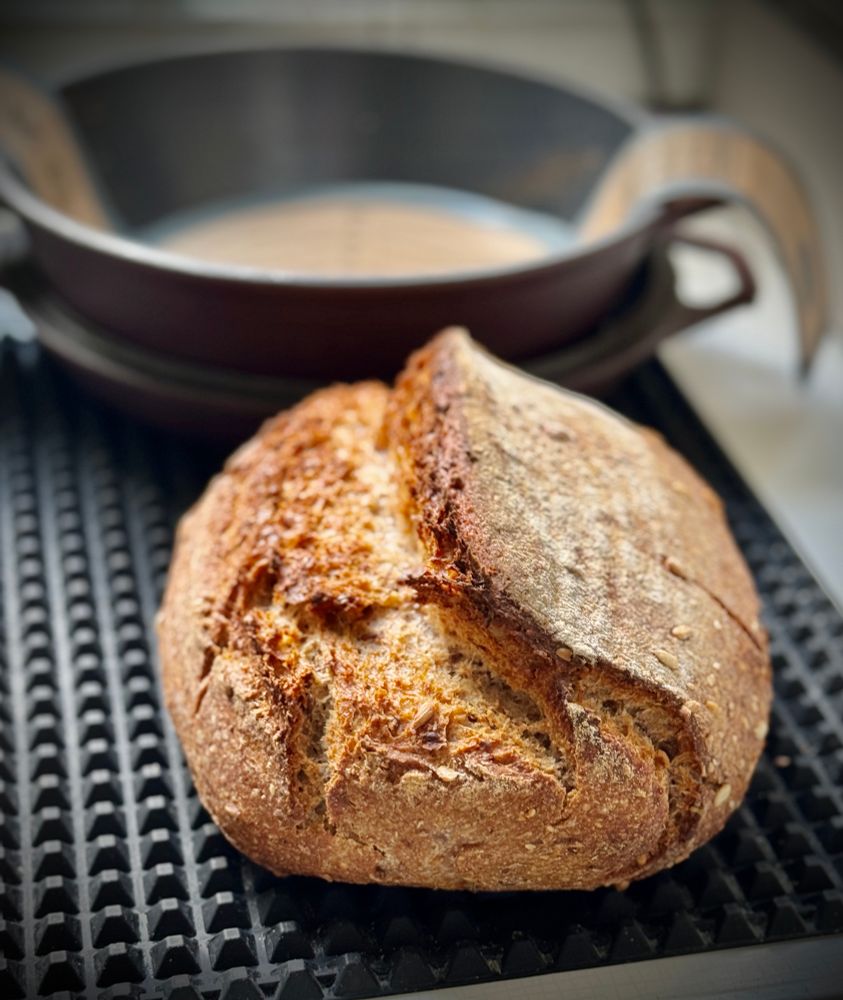 Das Foto zeigt ein frisch gebackenes, rundes Bauernbrot mit knuspriger, goldbrauner Kruste und kräftigem Ausbund (aufgerissene Stelle an der Oberfläche). Es liegt auf einer schwarzen Gummimatte, im Hintergrund steht ein offener, gusseiserner Topf, in dem das Brot vermutlich gebacken wurde.