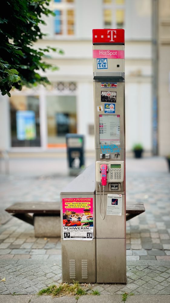 Auf dem Foto ist eine silberne Telefonzelle der Telekom zu sehen – schlank, modern und ohne Kabine. Oben leuchtet das rote T-Logo. Darunter steht „HotSpot“, denn hier gibt es auch WLAN. In der Mitte hängt ein graues Tastenfeld mit einem pinken Telefonhörer. Auf der ganzen Säule kleben bunte Tickets, Texte und Sticker. Unten ist ein Plakat für eine Veranstaltung mit Trampolinen und Tieren. Die Telefonzelle steht auf einem Trottoir (Gehweg) in einer Stadt mit Bäumen und Türen im Hintergrund.