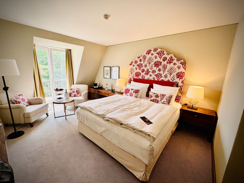 Das Foto zeigt ein helles, gemütlich eingerichtetes Hotelzimmer mit einem großen Doppelbett in der Mitte. Das Bett hat ein auffälliges rotes Kopfteil mit weiß-rotem Muster, das an Korallen erinnert, und ist mit mehreren Kissen dekoriert. Links neben dem Bett stehen zwei bequeme Sessel mit einem kleinen runden Tisch dazwischen, auf dem eine Etagere mit Süßigkeiten oder Obst steht. Die Wand hinter dem Bett ist cremefarben, und an beiden Seiten des Bettes stehen Nachttische mit Lampen. Das Zimmer hat einen großen Fensterbereich mit Vorhängen, durch den viel Tageslicht hereinkommt.