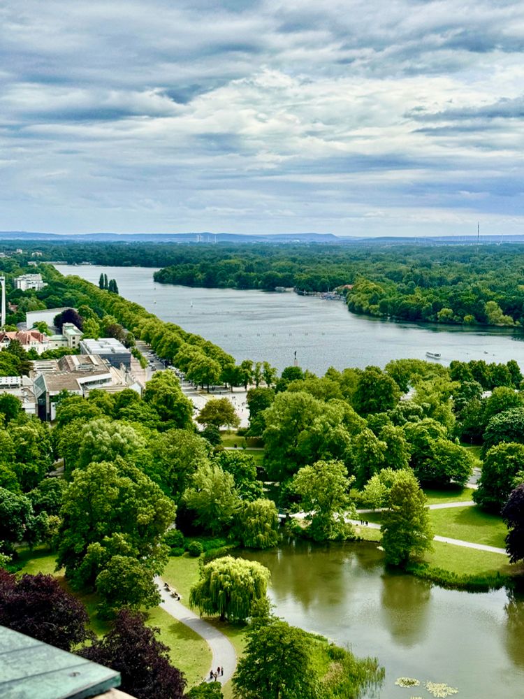 Im Vordergrund des Farbfotos liegt der idyllische Maschteich in Hannover, umgeben von dichtem, grünem Baumbestand. Der Teich ist ruhig und spiegelt das satte Grün der umliegenden Bäume wider. Ein schmaler Fußweg schlängelt sich am Ufer entlang, auf dem einige Menschen spazieren gehen oder auf Bänken verweilen. Besonders auffällig ist eine große, hängende Trauerweide direkt am Wasser – ihr Blätterdach neigt sich sanft über die Teichoberfläche und verleiht der Szene eine fast märchenhafte Atmosphäre.

Hinter dem Maschteich öffnet sich der Blick auf den deutlich größeren Maschsee, einen langgestreckten, künstlich angelegten See. Er liegt eingebettet in ein grünes Band aus Bäumen, das sich bis zum Horizont zieht. Am linken Ufer des Sees erkennt man Teile der Stadt – Gebäude, Wege und Menschen, die die Promenade entlanggehen. Auf dem Wasser sind kleine Boote zu sehen, die über die glatte Oberfläche gleiten.

Der Himmel darüber ist wolkig, mit hellen und dunklen Wolkenschichten, die dem Bild eine leicht dramatische, aber dennoch friedliche Stimmung verleihen. Die Landschaft wirkt weit und offen, voller Grün und Wasser – ein Ort der Ruhe mitten in der Stadt.