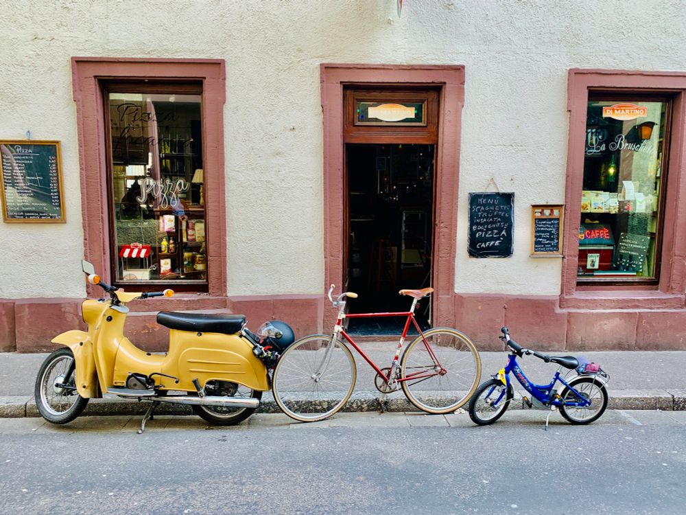 Das Foto zeigt drei verschiedene Zweiräder, die nebeneinander am Straßenrand vor einem kleinen Restaurant stehen. Ganz links steht ein altmodischer, gelber Motorroller. In der Mitte parkt ein klassisches, rotes Herrenfahrrad mit dünnen Reifen und einem braunen Sattel. Ganz rechts steht ein kleines, buntes Kinderfahrrad – blau mit gelben und roten Akzenten – mit Stützrädern und einem kleinen rosa Täschchen auf dem Gepäckträger.

Im Hintergrund sieht man die Fassade eines italienischen Lokals mit heller Wand und rötlich-braunen Fensterrahmen. An den Fenstern hängen Speisekarten, und durch das Glas erkennt man Regale mit italienischen Produkten. Die Tafel neben der Tür kündigt typische Gerichte wie Spaghetti, Pizza und Caffè an.

Die Szene wirkt ruhig und charmant – als würde sie zu einer kleinen Fahrradtour durch eine gemütliche Altstadt einladen. Die drei Zweiräder, vom Kinderrad bis zum Moped, erzählen eine kleine Geschichte von Mobilität und Generationen.