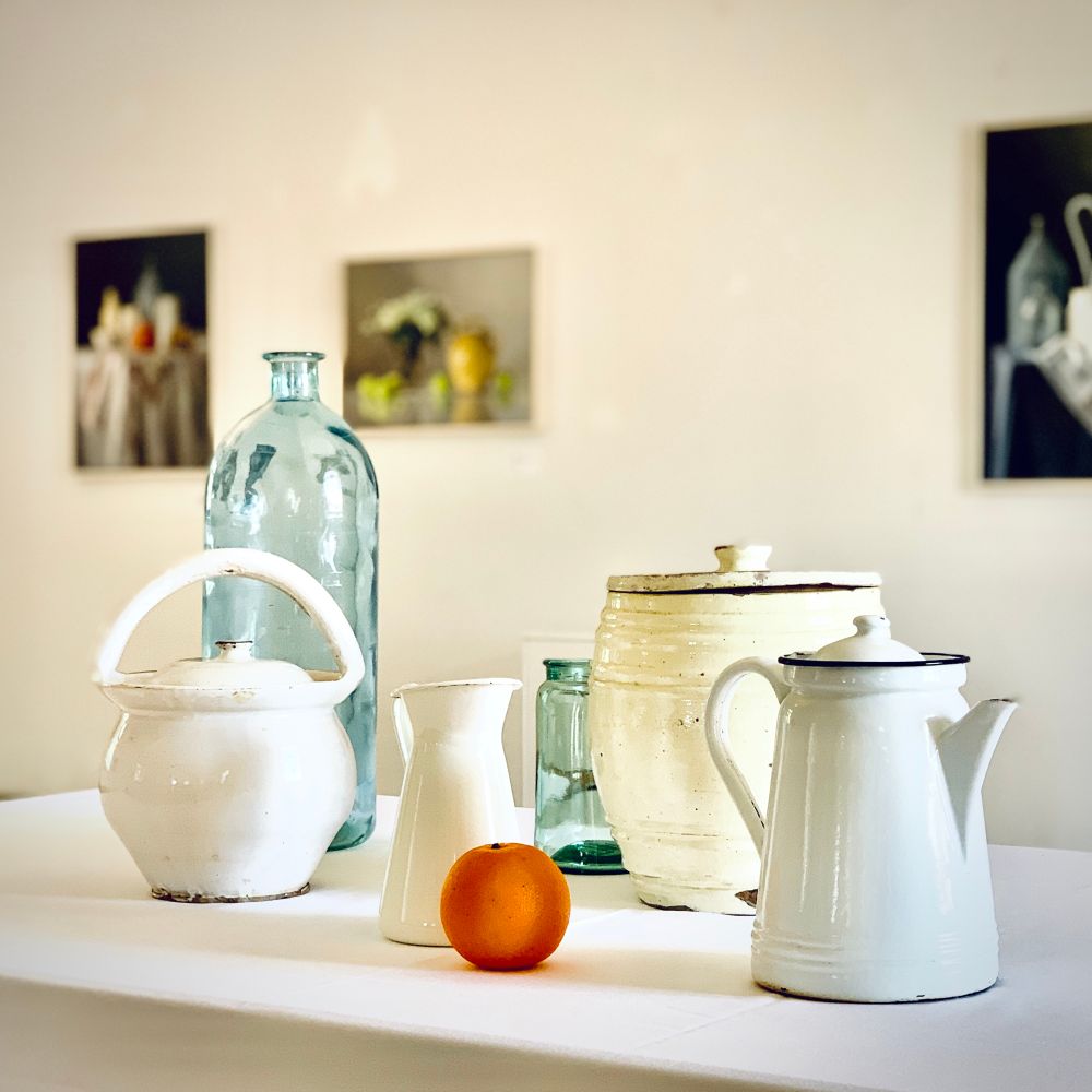 Das Foto zeigt ein schlichtes Stillleben auf weißem Tisch: alte Emaille-Kannen, Glasflaschen, ein Krug und eine Orange. Alles ist in gedeckten Farben gehalten, nur die Orange setzt einen leuchtenden Akzent. Der Hintergrund ist hell und ruhig, mit unscharfen Bildern an der Wand. Wenige Objekte, viel Raum – Minimalismus in Reinform.